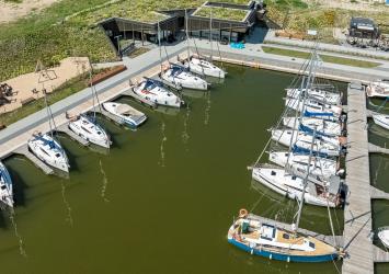 Na pierwszym planie basen portowy Przystani Nautica. Łodize zacumowane przy odnogach cumowniczych. W dalszym planie budynek kapitanatu i wypożyczalni rowerów.