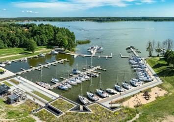 Widok na Przystań Nautica z lotu ptaka. Na pierwszy planie pawilon kapitanatu, budynek wbudowany w skarpę i przykryty trawą. Po prawej stronie keje czarterowe. Na środku basen portowy i wejście do portu. Z lewej u góry widać keje rezydenckie. W tle widać jezioro Święcajty 