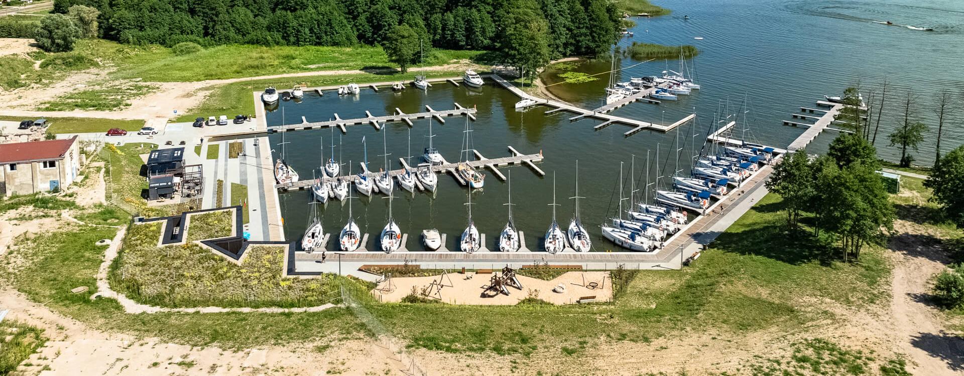 A bird's-eye view of Nautica Marina. In the foreground, the charter quay. On the left the captain's office, further to the left the old lumber buildings.  In the middle, the harbour basin and entrance to the harbour. Lake Swiecajty can be seen in the background on the right.