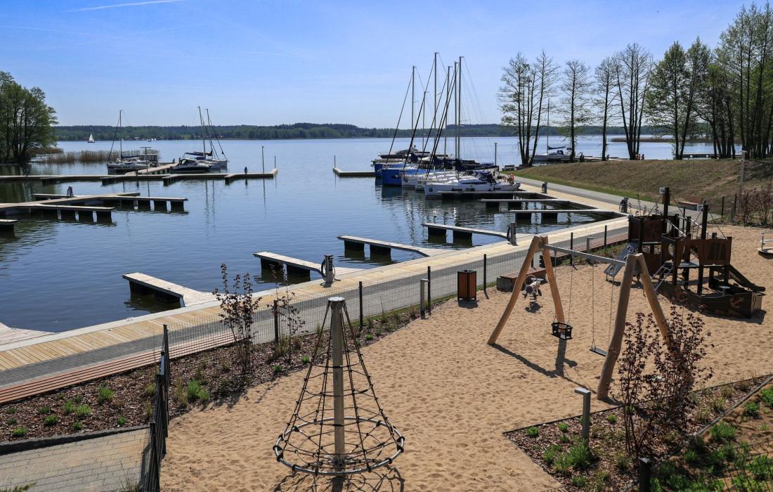 na pierwszym planie plac zabaw z piaszczystym podłożem, na drugim basen portu wewnętrznego. w oddali bezchmurne niebo i jezioro 