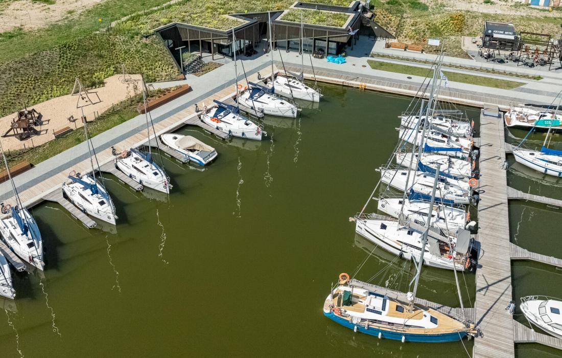 Na pierwszym planie basen portowy Przystani Nautica. Łodize zacumowane przy odnogach cumowniczych. W dalszym planie budynek kapitanatu i wypożyczalni rowerów.