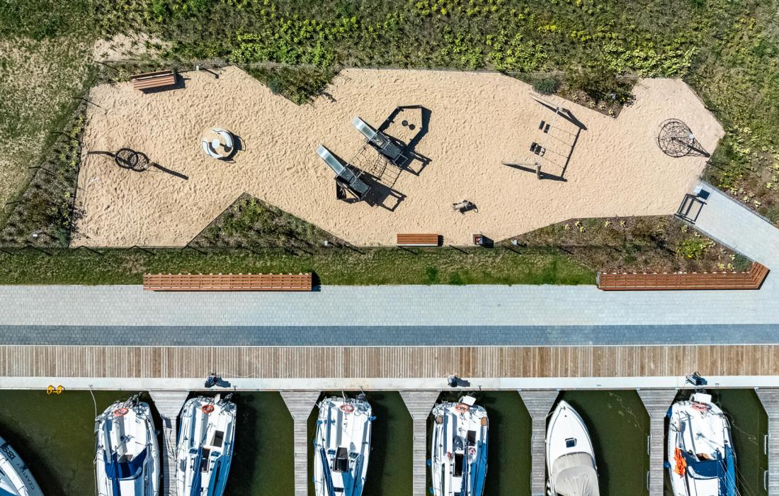 Widok na plac zabaw i nadbrzeże cumownicze z lotu ptaka. Widać plac zabaw i łodzie jachtowe zacumowane w porcie. 