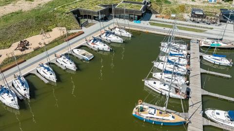 Na pierwszym planie basen portowy Przystani Nautica. Łodize zacumowane przy odnogach cumowniczych. W dalszym planie budynek kapitanatu i wypożyczalni rowerów.