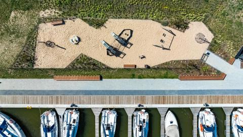Widok na plac zabaw i nadbrzeże cumownicze z lotu ptaka. Widać plac zabaw i łodzie jachtowe zacumowane w porcie. 