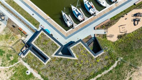 Na pierwszym planie budynek kapitanatu z lotu ptaka. Zarysowują się geometryczne kształty nieregularnej bryły budynku wbudowanego w skarpę i przykrytego trawą. Przy nadbrzeżu przycumowane łodzie żaglowe.