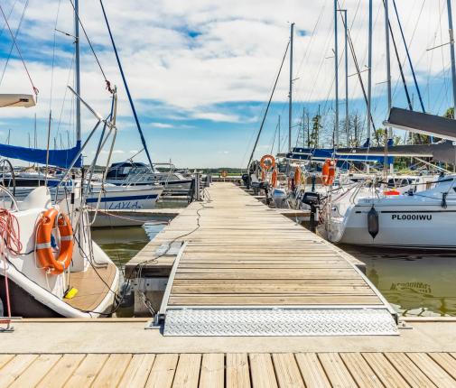 Pomost z odnogami cumowniczymi. Przy y-boomach przycumowane łódki. W tle błękitne niebo z białymi chmurkami.